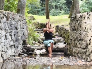 Kobieta w czarnej bluzce i niebieskich spodniach wykonująca pozycję jogi na kamieniach w lesie.