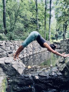 Osoba wykonuje mostek jogiczny nad wąskim korytem z wodą, otoczona zielenią drzew i kamiennymi murkami; plenerowa sesja jogi Iyengara w Bielsku-Białej.